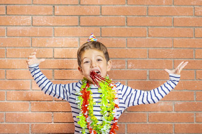 Portrait of excited boy wearing party equipment against brick wall