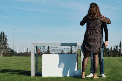 Couple standing by goal post on soccer field against blue sky