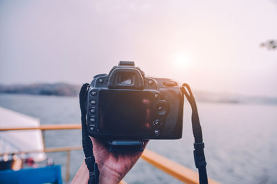 Cropped hand holding camera by lake during sunset