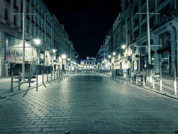 Empty footpath by illuminated street lights amidst buildings