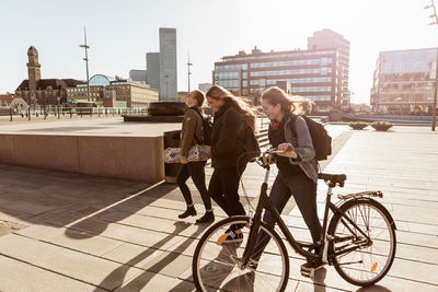 Teenage girl holding bicycle while walking with friends on pedestrian zone in city