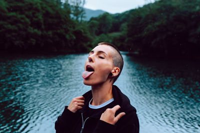 Short haired female with piercing wearing black hoodie looking up while catching raindrops with tongue near green forest and pond