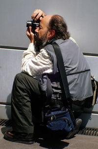 Full length of photographer with camera crouching on sidewalk by wall