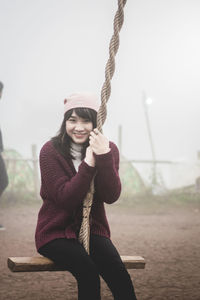 Portrait of young woman sitting on swing during foggy weather