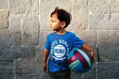 Thoughtful boy holding ball while standing against wall