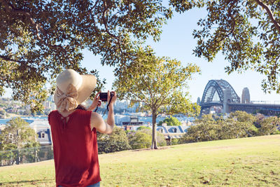 Rear view of woman photographing bridge in city against sky