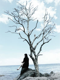 Woman sitting by bare tree on rock against sky
