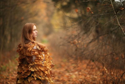 Thoughtful young woman covered with leaves standing at forest during autumn