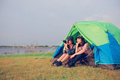 Smiling woman sitting by friend photographing while sitting in tent on land by lake