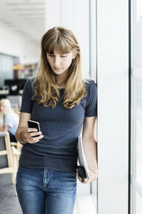 Young female student using smart phone while leaning on window at library