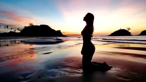 Silhouette woman kneeling at beach against sky during sunset