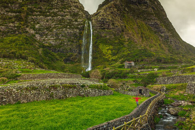 Woman arms outstretched walking on stone wall with waterfall and mountain background