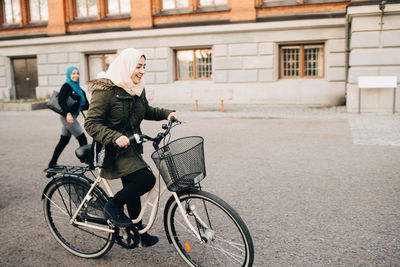 Happy young muslim woman cycling on street with friend walking in city