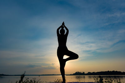 Silhouette woman exercising at lakeshore against sky during sunset