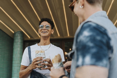 Happy young man in sunglasses holding wrap sandwich and standing with male friend
