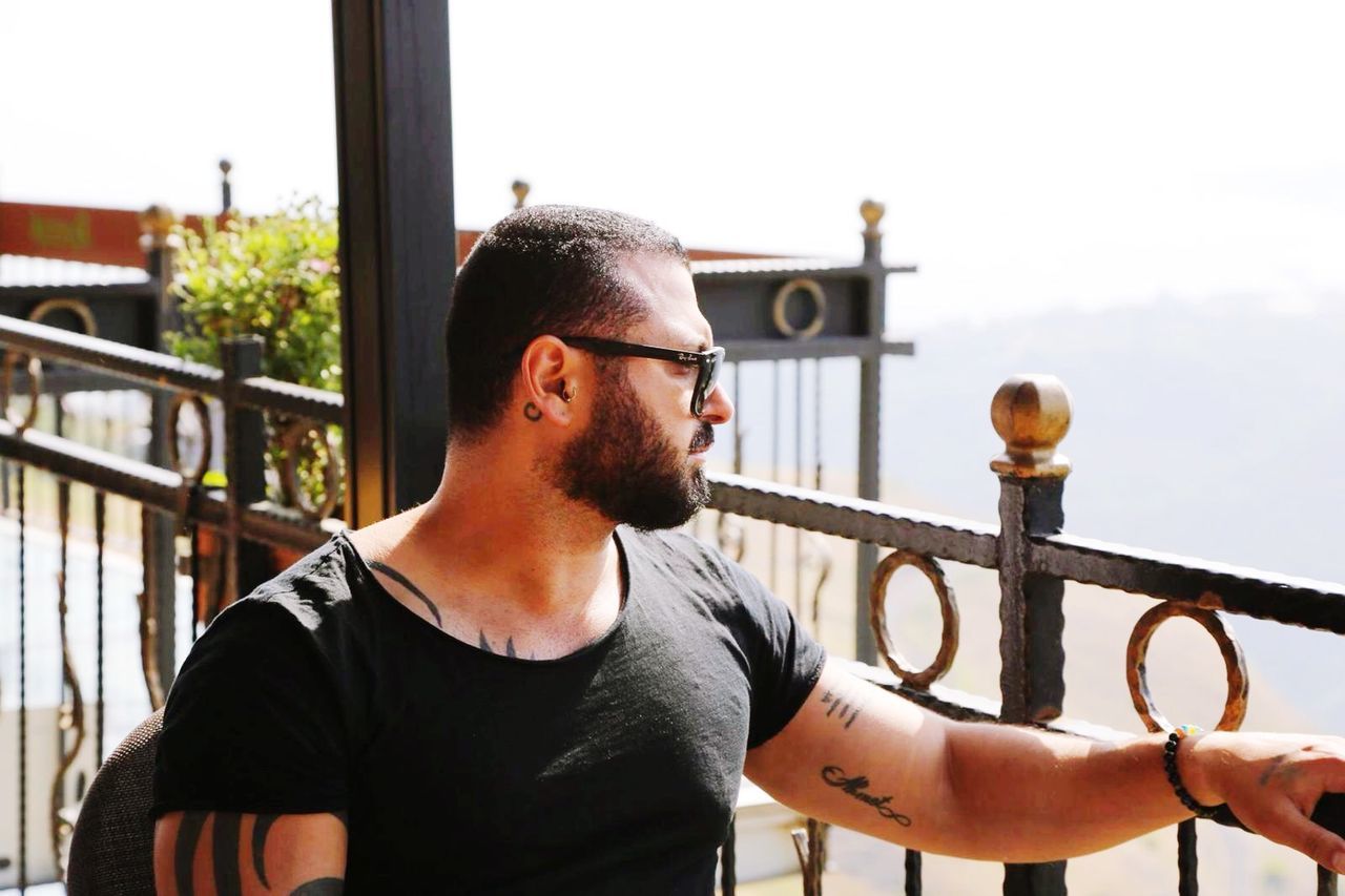 one person, railing, lifestyles, real people, beard, casual clothing, waist up, facial hair, day, glasses, standing, architecture, young adult, leisure activity, men, focus on foreground, portrait, mid adult, outdoors, hairstyle, contemplation