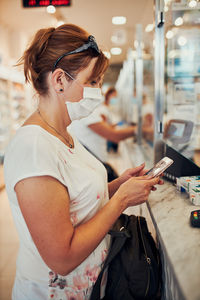 Side view of woman wearing mask using mobile phone at supermarket