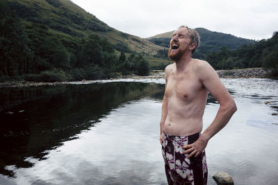 Shirtless laughing man standing at lake against sky