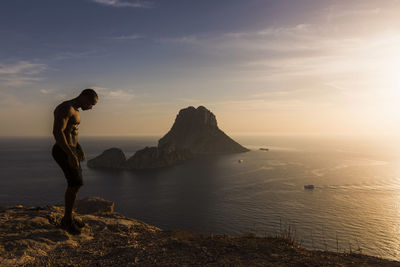 Side view of muscular man standing on rock by sea against sky during sunset