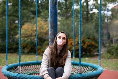 Smiling young woman blowing bubblegum while sitting in swing at playground