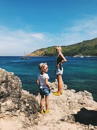 Full length of mother and daughter standing on rock formation at beach against sky