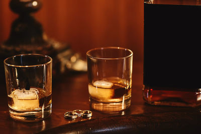 Two glasses of cognac with ice, bottle with mockup and gold wedding rings on a wooden table. wedding