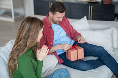 Intrigued merry man unbracing red ribbon on giftbox sitting on sofa next to woman presenting gift