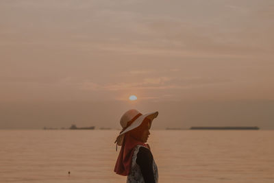 Rear view of woman standing at beach against sky during sunset