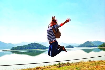 Portrait of happy young woman jumping by lake against clear sky during sunny day