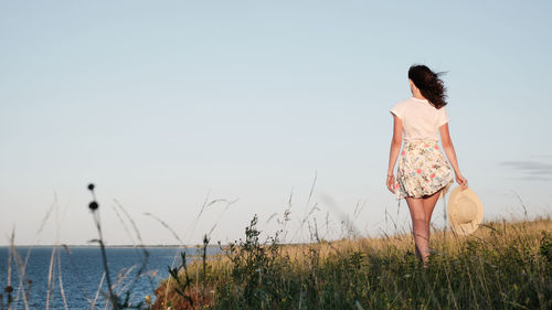 A woman walks along the shore and holds a hat in her hand.