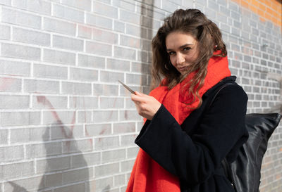 Portrait of young woman holding smart phone while standing against wall