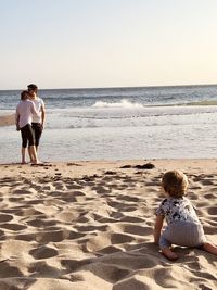 Baby boy looking at couple embracing at beach against clear sky