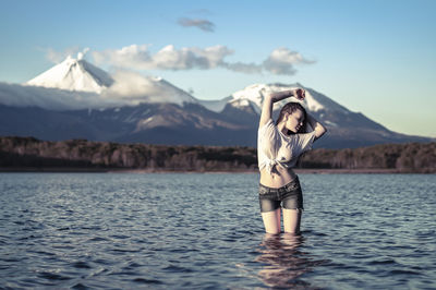 Young woman with arms raised standing in lake against snowcapped mountain