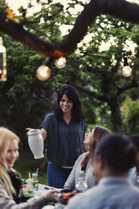 Smiling woman holding jug while friends sitting at dining table during party