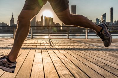 Low section of man running on boardwalk against buildings during sunrise in city