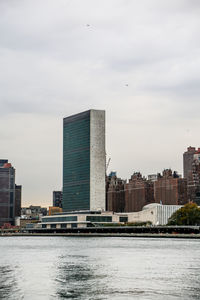 Modern buildings by river against cloudy sky in city