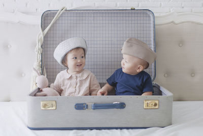 Cute girl and boy sitting in suitcase on bed at home