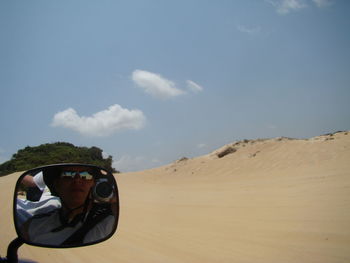 Reflection of man on side-view mirror on vehicle at desert against sky