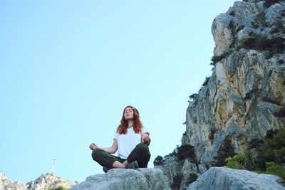 Woman sitting in lotus position on rock against clear sky