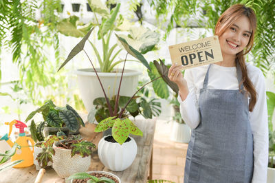 A woman with a smiling face in an apron stands holding a welcome open sign in the green house.