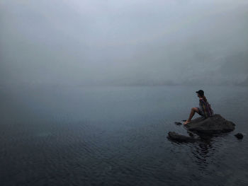 Side view of woman sitting on rock in lake during foggy weather