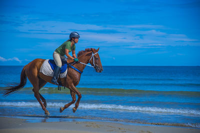 Full length of woman riding horse at beach against blue sky