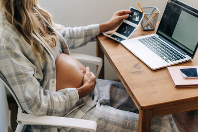 Unrecognizable pregnant woman sitting at the table looking at her baby's ultrasounds at home