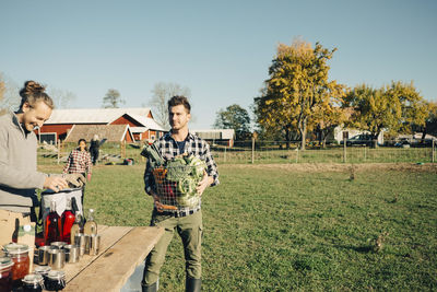 Male farmer arranging table while friend carrying basket of organic vegetable at field