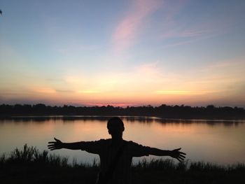 Silhouette man standing at lakeshore with arms outstretched against sky during sunset