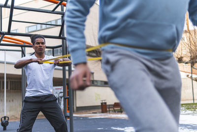 Young man holding stretching band while exercising with friend in city