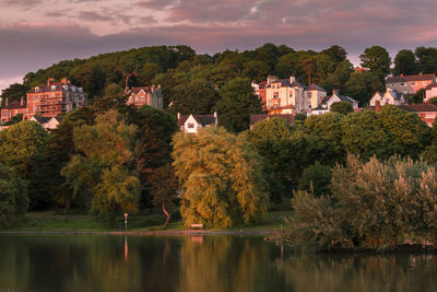 Trees by lake and buildings against sky during autumn