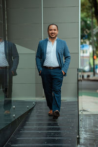 Happy hispanic mature male manager in suit holding hands in pockets and looking at camera with smile while walking near glass wall on sunny day on city street