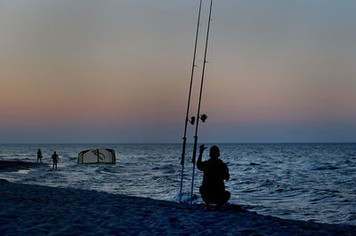 Silhouette people fishing on beach against sky during sunset