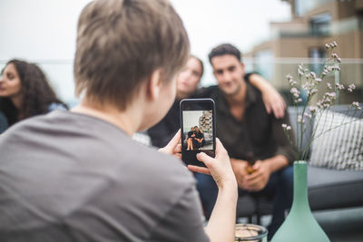 Rear view of woman photographing friends through smart phone during terrace party